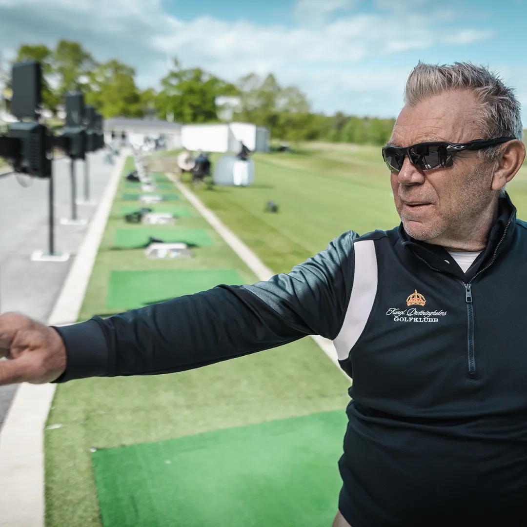 Man wearing sunglasses pointing at something on a golf practice range with equipment and greenery in the background.