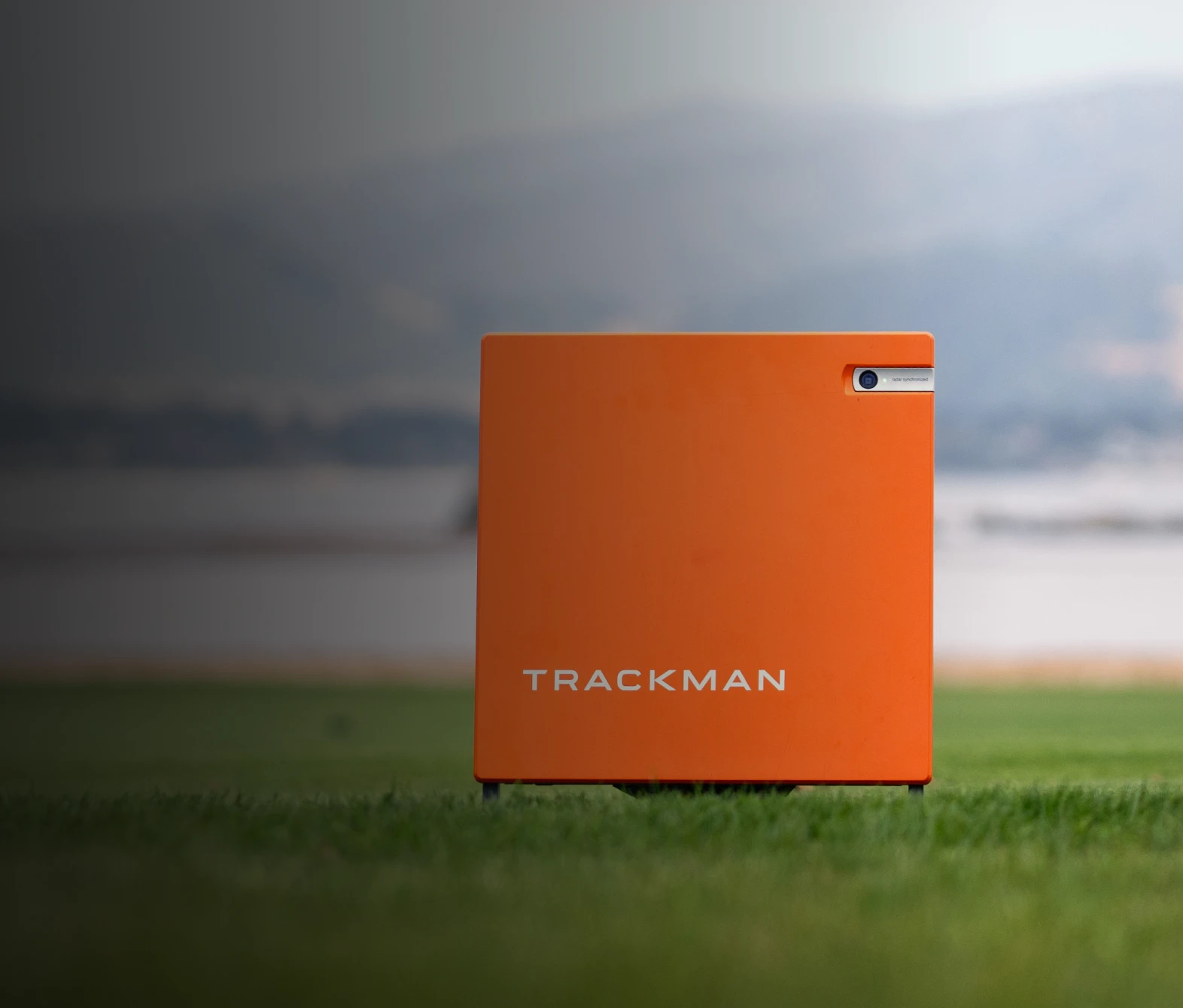 Orange Trackman device on grass with blurred mountains in the background.
