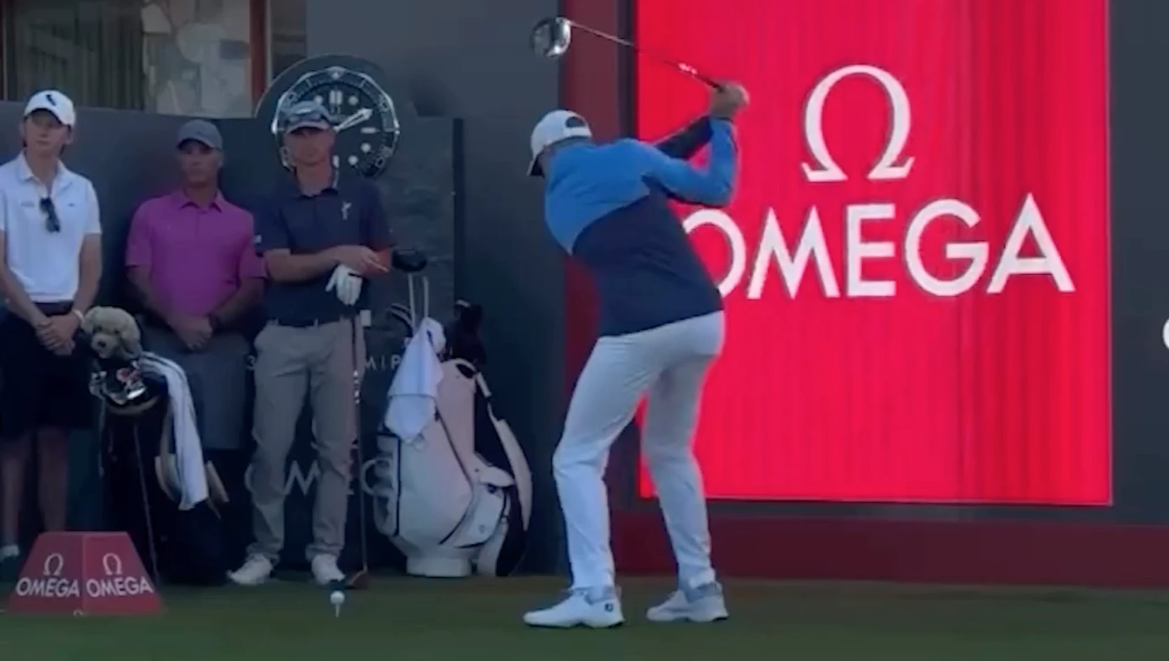 Golfer in blue swings club at tee; spectators stand nearby. Omega logo on large red display in background.