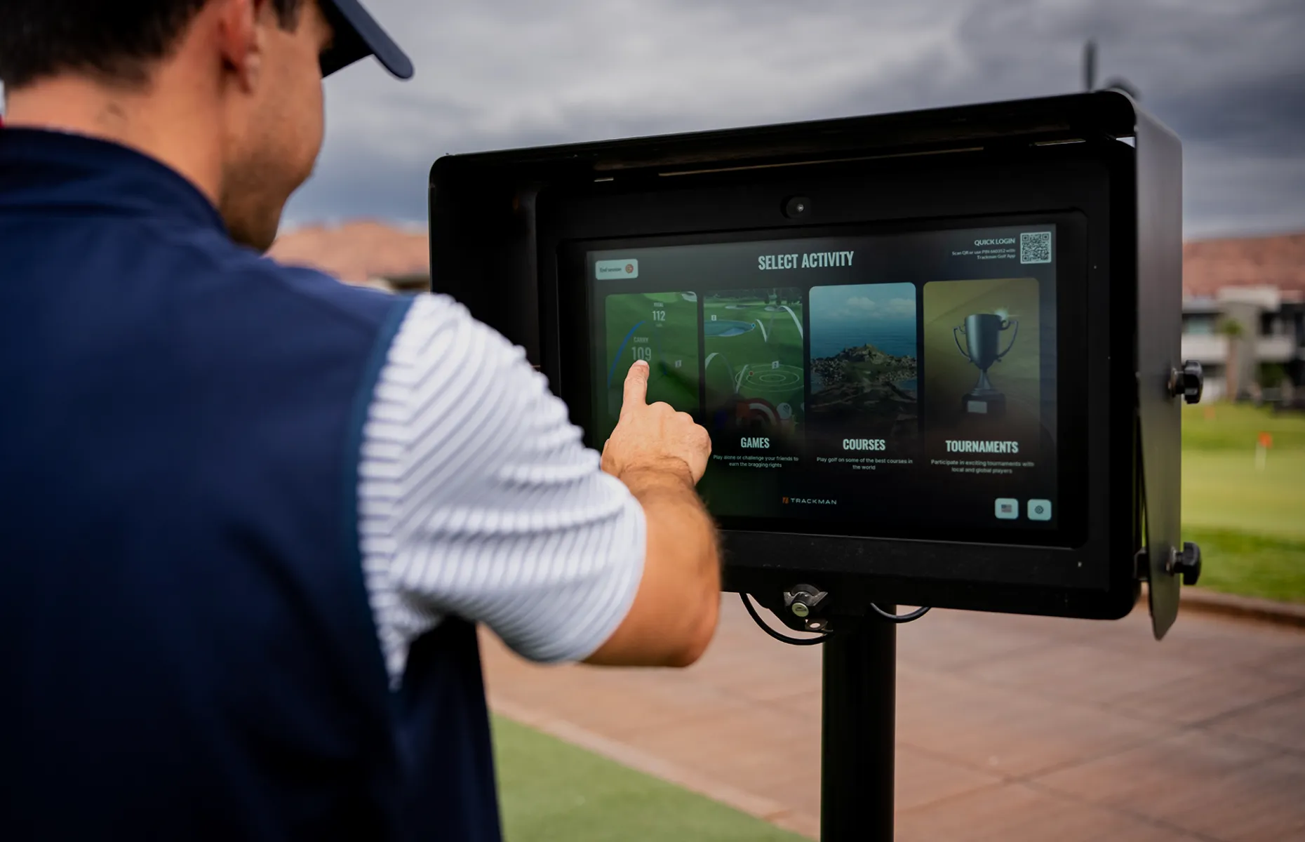 Person using a touchscreen outdoors, selecting activities like games and tournaments, with a golf course visible in the background.