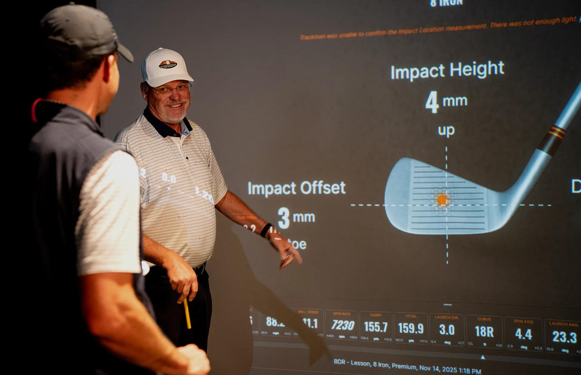 Two golfers in a golf simulator room discuss data on a screen, showing impact height, offset, and other statistics with a golf club diagram.