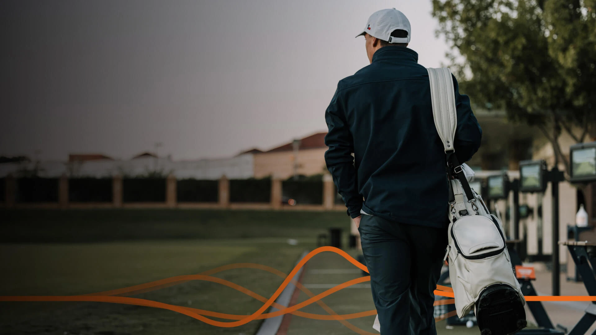 A person in a dark jacket and cap walks on a golf course carrying a golf bag. The background features trees and a row of golf carts.