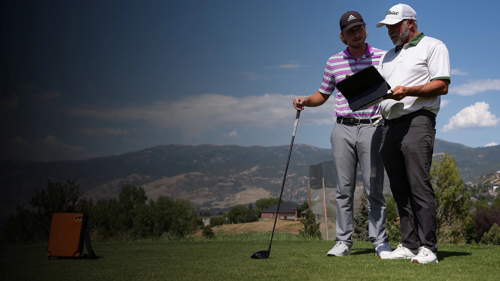 Two golfers stand on a lush course, one holding a club and the other a tablet, with mountains and a partly cloudy sky in the background.