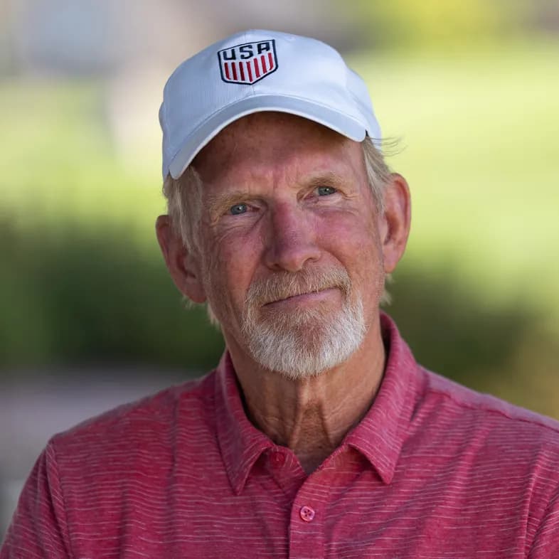 Elderly man with a white beard wearing a white USA cap and red polo shirt, outdoors with a blurred green background.