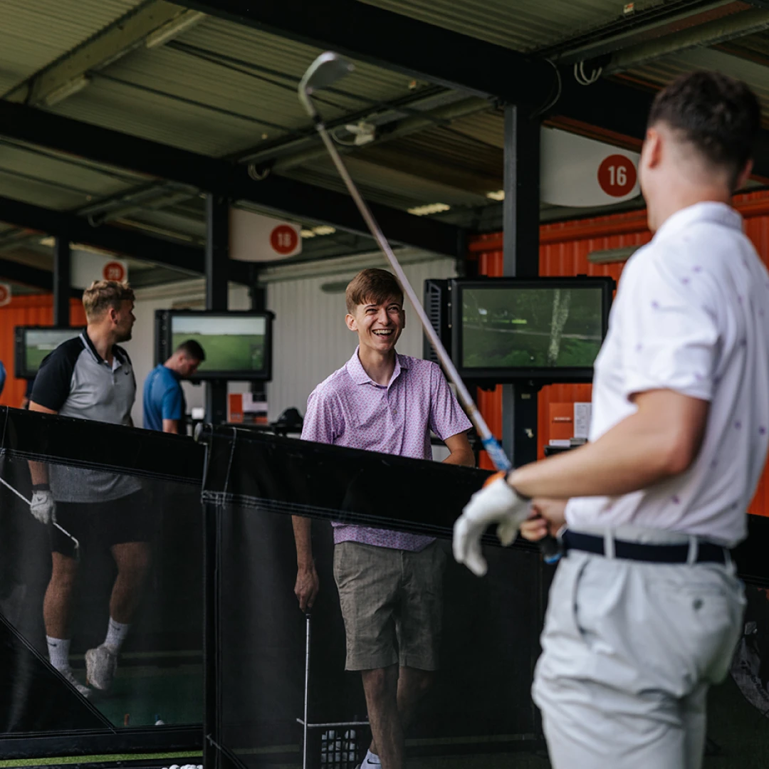 Three men enjoying golf at an indoor driving range, with one man playfully swinging a club and another laughing enthusiastically.