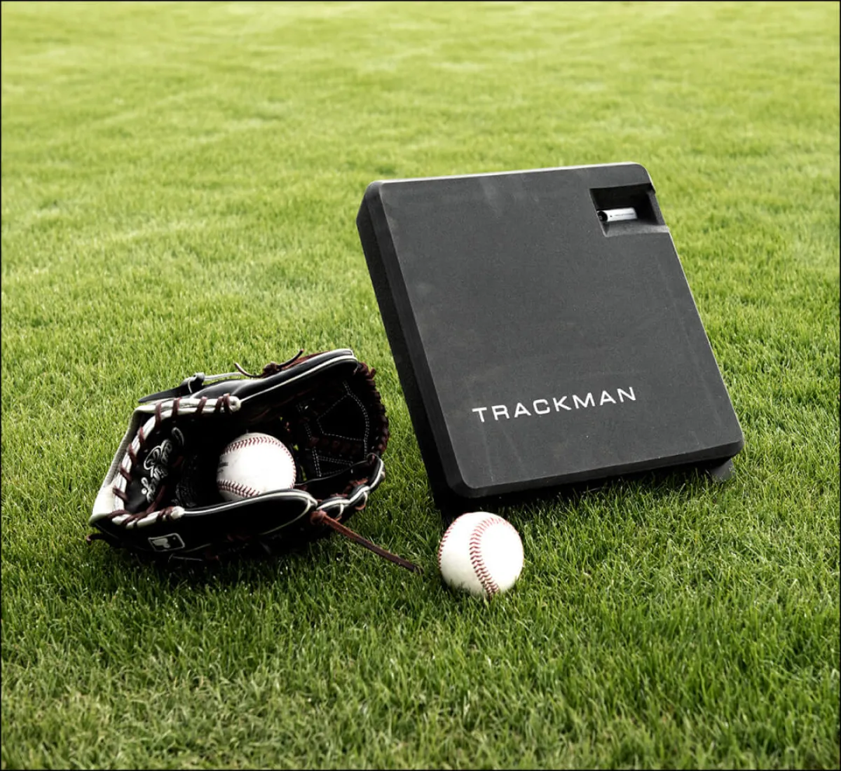 A baseball glove with a ball inside sits on grass next to a Trackman device and another baseball.