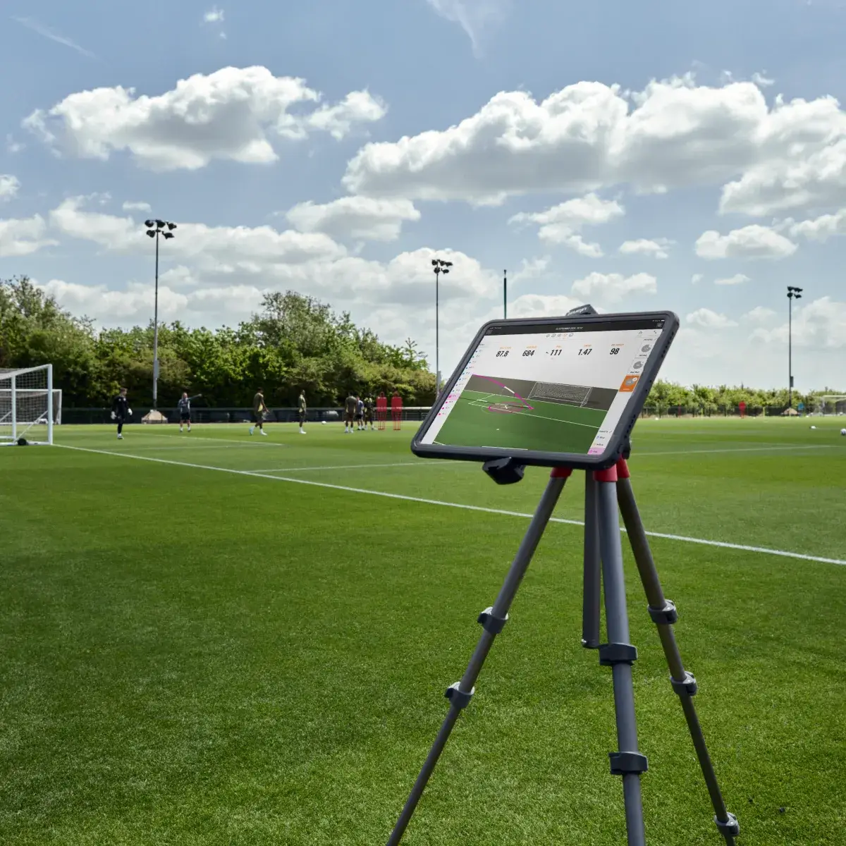 A tablet on a tripod stands on a soccer field, displaying data. Goalposts and players are visible in the background under a partly cloudy sky.