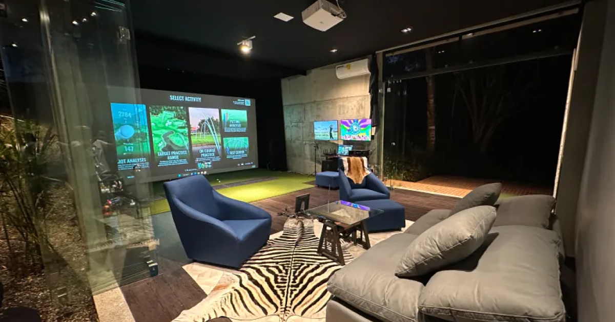 A Trackman indoor home golf simulator with a large screen, blue chairs, gray sofa, zebra rug, and glass walls overlooking a garden.