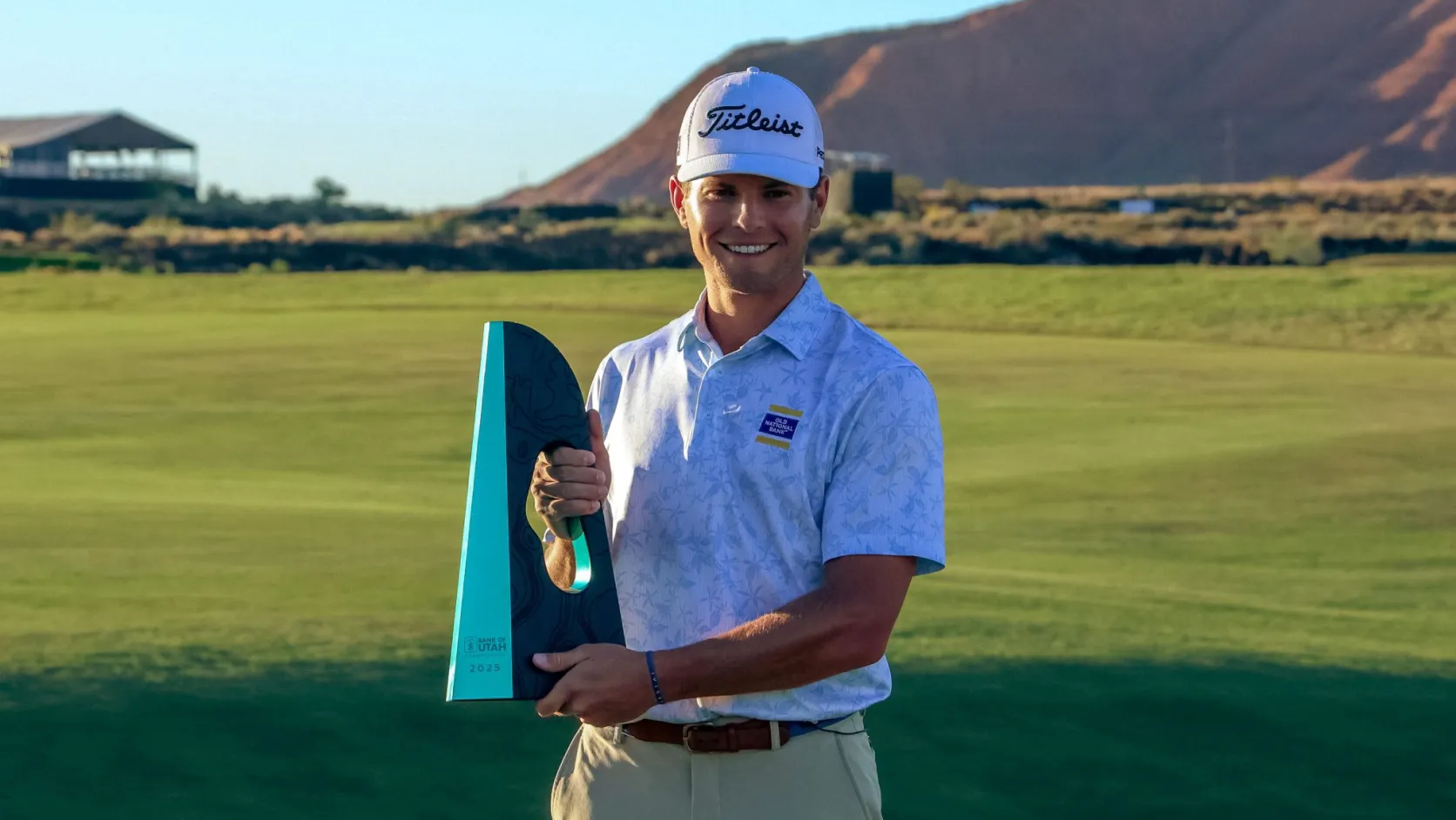 Michael Brennan smiling and holding a trophy on a golf course, wearing a white cap and shirt with a mountain in the background.