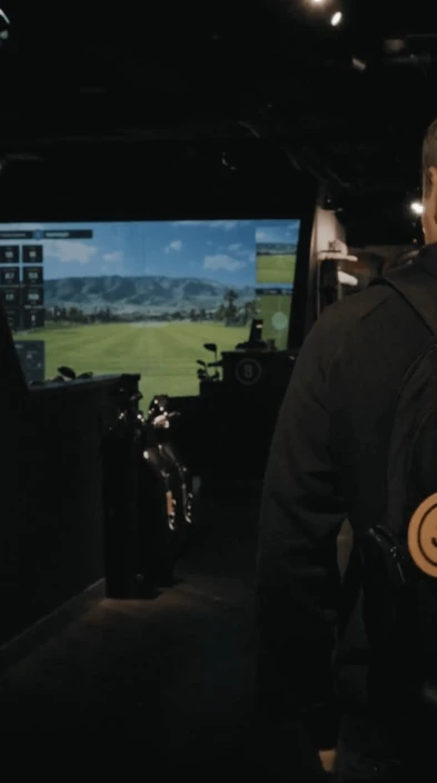 Person in a dark room with a golf simulator displaying a scenic course on a large screen. Golf bags are visible on the side.