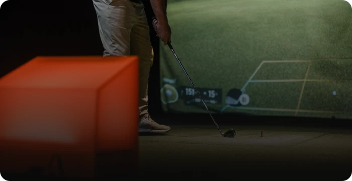 Golfer prepares to swing on an indoor virtual golf simulator, with a large glowing cube in the foreground.