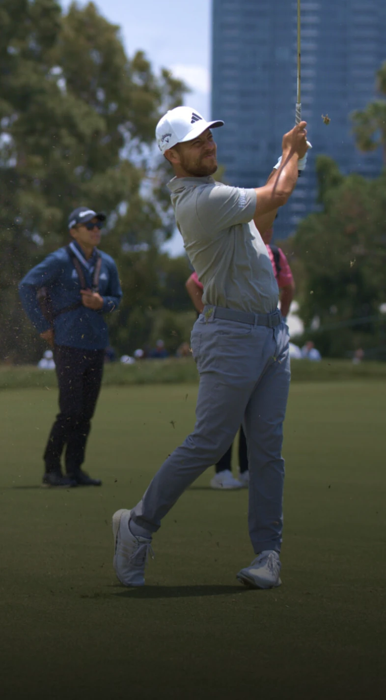 Golfer in mid-swing on a grassy course, with spectators in the background and trees nearby.