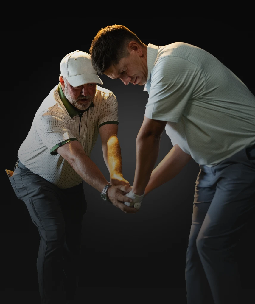 Two men focused on perfecting a golf swing, one guiding the other's grip on the club, against a dark background.